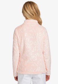 Chaqueta de forro polar rosa claro con textura floral sutil y cuello. Presenta mangas largas y un dobladillo recto. Sin cierres ni adornos visibles.