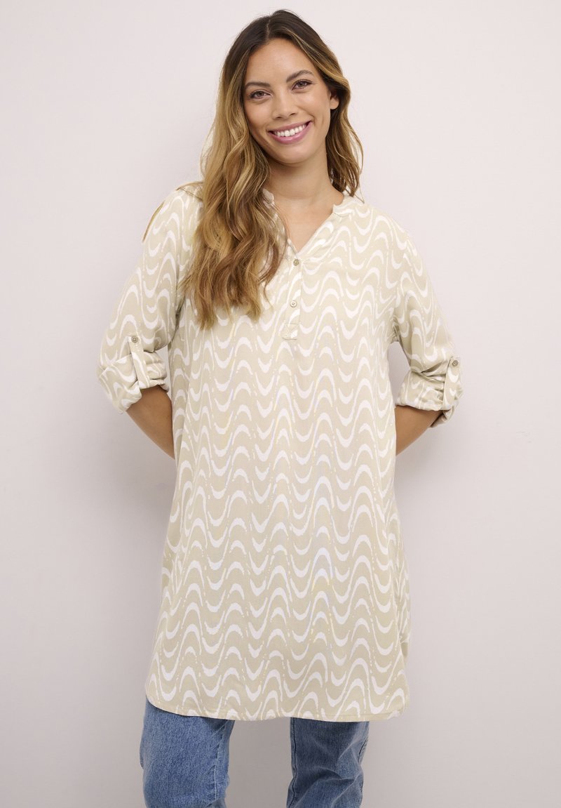 Beige tuniek met een golvenpatroon in het wit, voorzien van een knoopsluiting, omgeslagen mouwen en een losse pasvorm. Zachte stoftextuur.