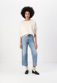 Überdimensioniertes cremefarbenes Sweatshirt mit gesprenkelter Textur, kombiniert mit hoch taillierten, hellblauen, weiten Jeans und schwarzen Mokassins.