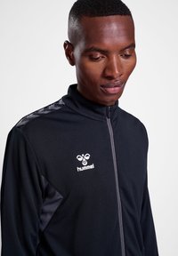 Schwarze Sportjacke mit einem Reißverschlusskragen, aus strukturiertem Stoff und grauen Akzenten an den Schultern. Hummel-Logo auf der Brust.