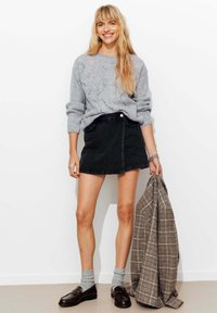 Femme souriante avec de longs cheveux blonds, portant un pull gris, une jupe noire, des chaussettes grises et des mocassins marron, tenant une veste à carreaux à ses côtés.