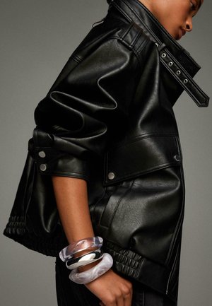Veste en cuir noir avec un col haut, poches latérales et détails en métal argenté. Modèle portant des bracelets transparents et argentés au poignet.