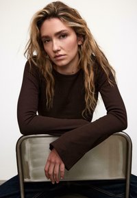 Top marrone a maniche lunghe con costine e polsini ampi, indossato da una persona seduta su una sedia. Lo sfondo è neutro, mettendo in risalto la texture del top.