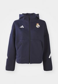 Marinblå zip-up jacka med huva, med vita Adidas-ränder och Real Madrid-logotyp. Tillverkad av mjukt, strukturerat tyg. Inkluderar framfickor.