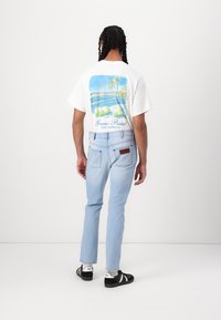 Lichtblauwe jeans met een slim fit, gecombineerd met een wit T-shirt met een palmboom en strandontwerp in blauw en groentinten, en zwarte sneakers.
