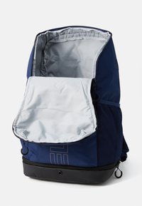 Sac à dos bleu marine avec un intérieur gris clair, doté d'une fermeture zippée, d'accents en maille et d'une base noire robuste. Comprend un rabat à l'avant.