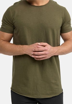 Homme portant une chemise à manches courtes vert olive, les bras pliés et les mains jointes au niveau de la taille.