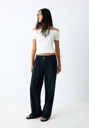 Žena v bielom topánku s odhalenými ramenami a širokými čiernymi nohavicami, stojaca a pozerajúca sa doľava na jednoduchom bielom pozadí.