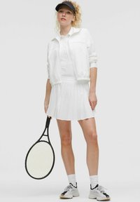 Robe de tennis plissée blanche avec une veste zippée, visière noire, chaussettes blanches et baskets noir et blanc, tenant une raquette de tennis noire.