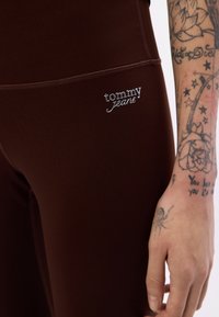 Brązowe elastyczne legginsy z pionowymi szwami i srebrnym logo "tommy jeans" na udzie. Gładka faktura, dopasowany krój, odpowiednie do codziennego noszenia.