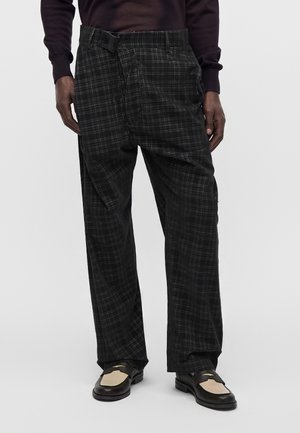 Homme portant un pantalon ample à carreaux noirs et gris avec un détail de bouton sur le côté, associé à des mocassins noirs et beiges.