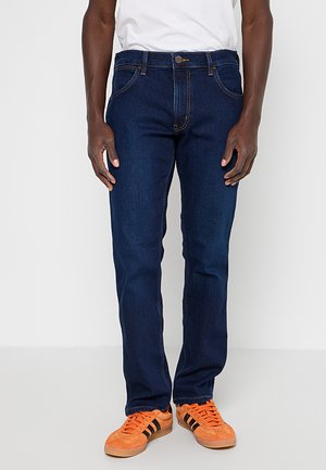 Mężczyzna noszący ciemnoniebieskie proste jeansy, pomarańczowe sneakersy i białą koszulę, stojący na tle jednolitego jasnego tła.