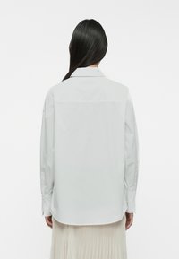 Camisa blanca de mangas largas con una canesú en la parte posterior, puños abotonados y una textura suave y lisa. Se pueden ver los puños en forma de campana y un dobladillo de camisa.