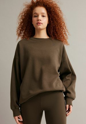 Ung kvinde med krøllet rødt hår iført en oversized olivengrøn sweatshirt og matchende tætsiddende bukser, stående foran en neutral baggrund.