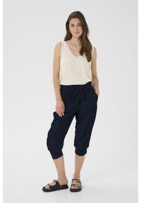 Pantaloni jogger blu con vita elastica e coulisse, abbinati a un top senza maniche beige chiaro, indossati con sandali neri con dettagli dorati.