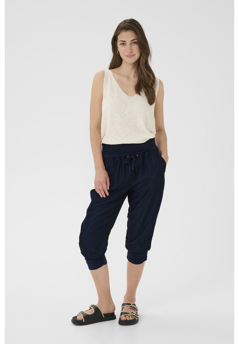 Pantaloni jogger blu con vita elastica e coulisse, abbinati a un top senza maniche beige chiaro, indossati con sandali neri con dettagli dorati.