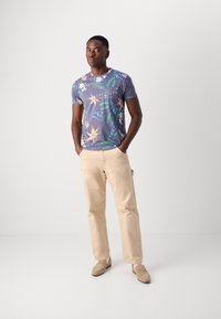 Camisa de manga corta azul marino con estampado floral que presenta flores coloridas y hojas verdes. Combinada con pantalones beige y mocasines marrón claro.