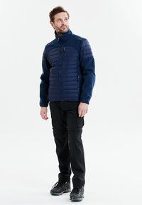 Marineblå quiltet jakke med høj krave og lynlåslommer, parret med sorte cargo bukser og grå vandresko. Enkel design, funktionelle elementer.