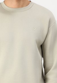 Felpa grigia chiara con collo a giro e maniche lunghe. Presenta un logo inciso e discreto sul lato sinistro del petto. Texture liscia.