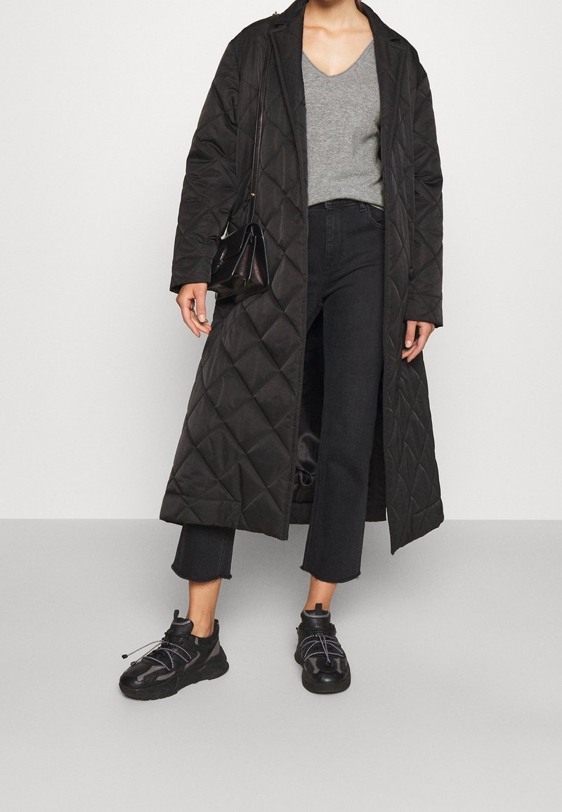 Personne portant un long manteau noir matelassé, un pull gris col V, un jean noir court, des baskets noires, et portant un petit sac noir à l'épaule.