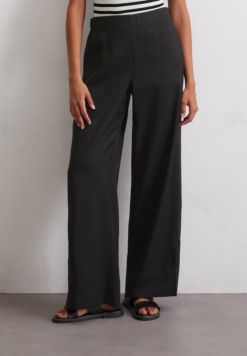 Pantalons larges noirs en tissu lisse avec une taille élastique, présentant une coupe décontractée qui descend jusqu'aux chevilles.