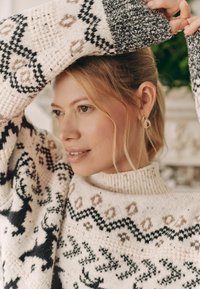Strickpullover mit hohem Kragen, cremefarbenem Hintergrund und schwarzen sowie erdigen Mustern. Strukturierter Stoff mit Rentiermotiven.