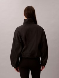 Black, lightweight jacket with a high collar, loose fit, and elastic cuffs. Features a cinched waist and discreet drawstring accents.
