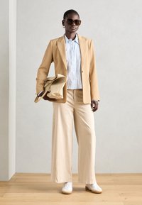 Béžový elegantný blejzer na svetlomodrej košeli na gombíky, krémové široké nohavice, biele tenisky a oversized slnečné okuliare s neutrálnou taškou.