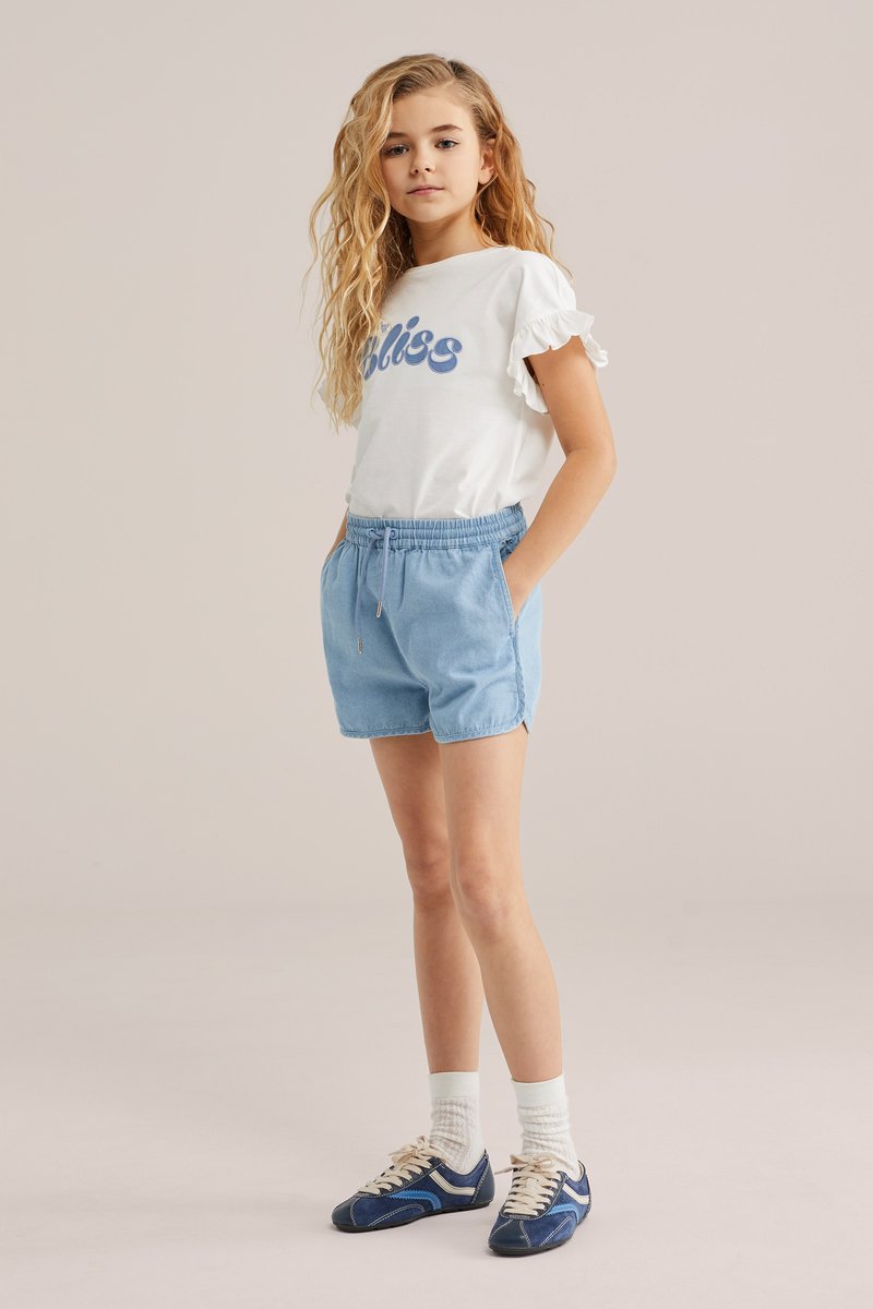 Plavuša s dugom kovrčavom kosom, odjevena u bijelu majicu s naboranim rukavima, svijetloplave kratke hlače, bijele čarape i plave tenisice, stoji s rukama u džepovima.