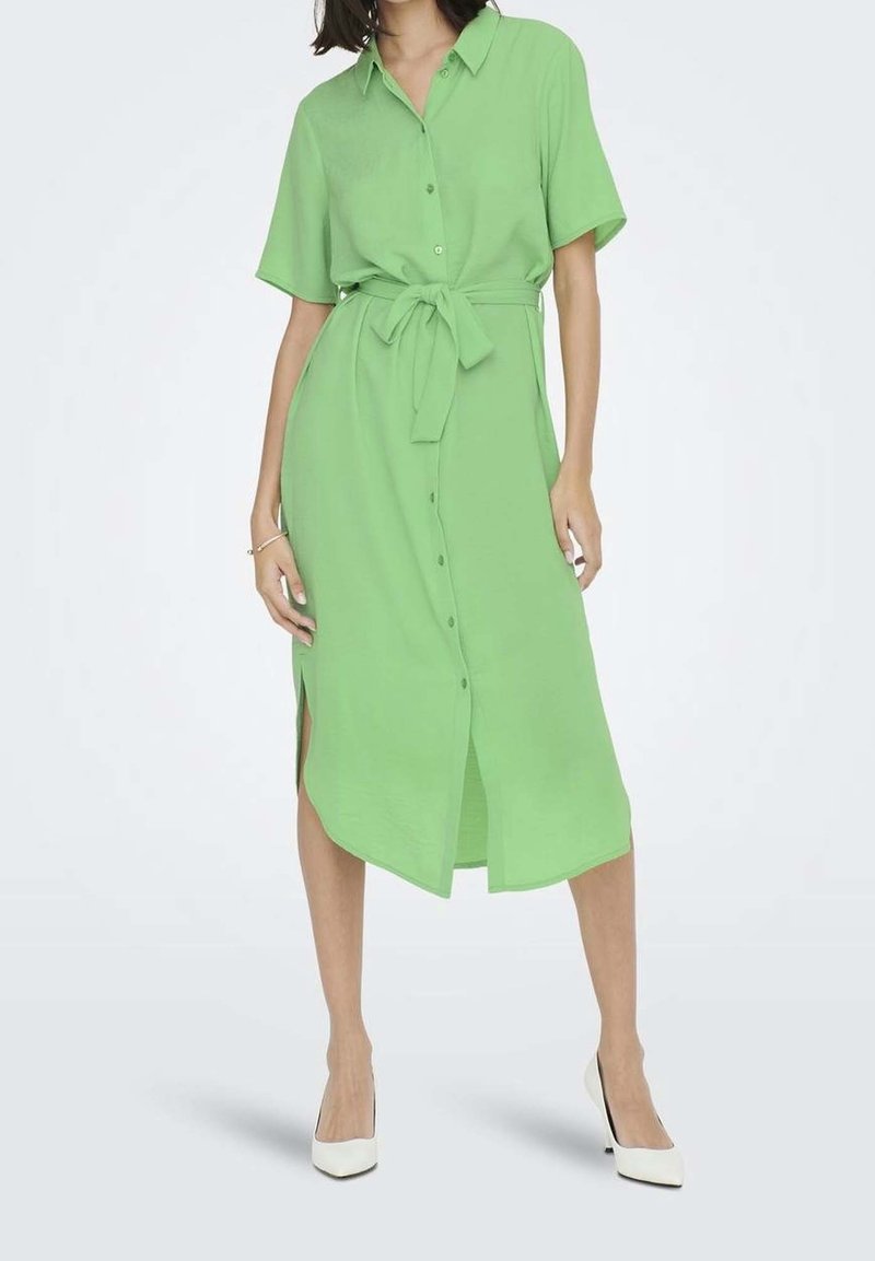 Femme portant une robe midi vert clair boutonnée à manches courtes avec une ceinture à la taille et des talons blancs, debout devant un arrière-plan uni.