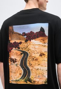 Μαύρο μπλουζάκι με πίσω γραφικό ενός στριφτού δρόμου στην έρημο, δύο μοτοσικλετιστές, πινακίδα "ΣΤΡΟΦΗ 15 MPH" και κόκκινα τριαντάφυλλα πάνω από το τοπίο.