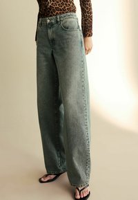 Vidjeans i urtvättad grön denim, med hög midja och klassisk femfickdesign. Kombineras med svarta flip-flops och en mönstrad topp.