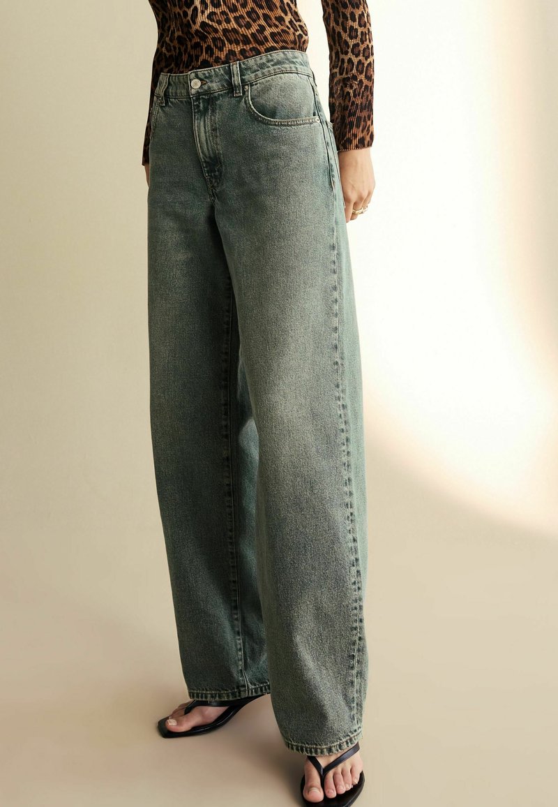 Vidjeans i urtvättad grön denim, med hög midja och klassisk femfickdesign. Kombineras med svarta flip-flops och en mönstrad topp.