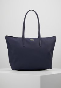 Sac fourre-tout bleu marine en tissu texturé avec deux longues anses, fermeture éclair et un petit logo vert en accent.