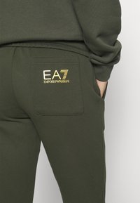 Detailní záběr na olivově zelené tepláky se zadní kapsou a zlatým logem "EA7 Emporio Armani" s rukou odpočívající v boční kapse.