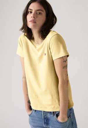 Giovane con capelli scuri e lunghi fino alle spalle, che indossa una maglietta giallo pallido e jeans blu, in piedi con le mani in tasca davanti a uno sfondo semplice.