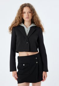 Zwarte cropped blazer met een enkele knoop, gecombineerd met een grijze hoodie en een zwarte rok met een voorvak en zilveren knopen.