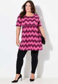 Růžová a černá tunika s chevronovým vzorem a krátkými rukávy, kombinovaná s černými legínami a klasickými černými vysokými podpatky.