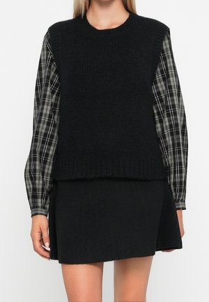 Femme portant un gilet en tricot noir sans manches par-dessus une chemise à manches longues à carreaux noirs et blancs et une jupe noire.