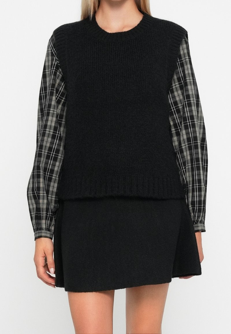Femme portant un gilet en tricot noir sans manches par-dessus une chemise à manches longues à carreaux noirs et blancs et une jupe noire.