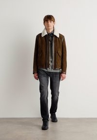 Chaqueta de ante marrón con cuello de borreguito blanco, cierre de cremallera en el frente y dos bolsillos laterales, combinada con jeans grises y zapatillas negras.
