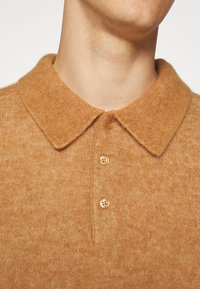 Beige stickad tröja med en mjuk textur, med en krage och tre knappar fram.