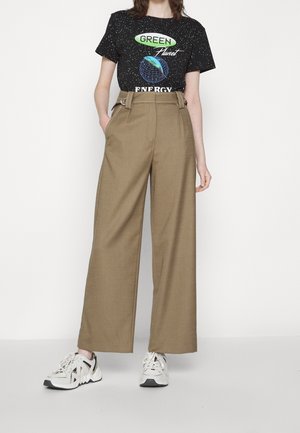 Persoon draagt een zwart t-shirt met de opdruk "Green Planet Energy", hoog getailleerde wijde bruine broek en witte sneakers, staande tegen een effen achtergrond.