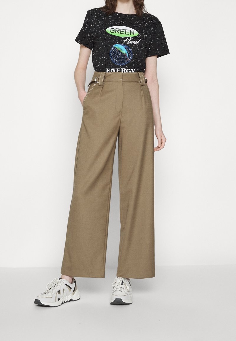 Personne portant un t-shirt noir « Green Planet Energy » avec un motif, un pantalon marron taille haute à jambes larges et des baskets blanches, debout devant un fond uni.