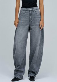 Jeans de denim gris de pierna ancha con cintura alta, que cuentan con bolsillos frontales, un cierre de botón y una textura desgastada. Un calzado liso y oscuro completa el look.