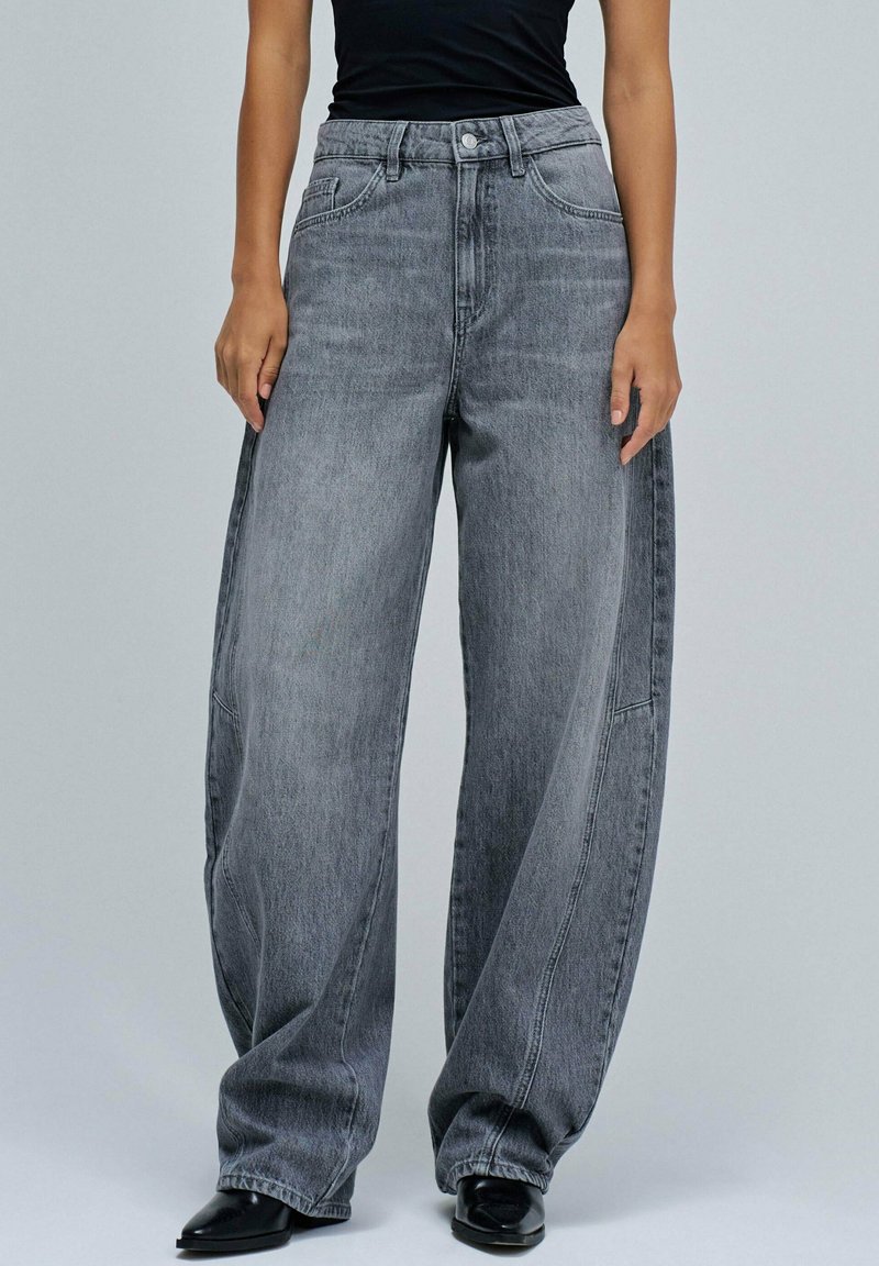 Jeans de denim gris de pierna ancha con cintura alta, que cuentan con bolsillos frontales, un cierre de botón y una textura desgastada. Un calzado liso y oscuro completa el look.