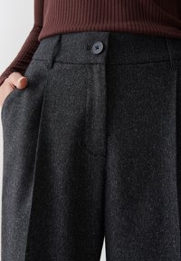 Personne portant un pantalon texturé gris foncé à taille haute, avec une fermeture à bouton et une main placée dans la poche gauche, associé à un haut marron à côtes.