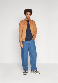 Giacca bomber in suede marrone con polsini a coste, abbinata a jeans blu larghi e sneakers multicolori. Sfondo neutro.