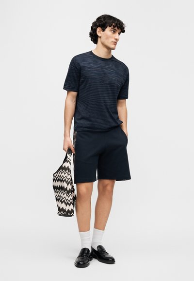 Μπλε-θαλάσσης ριγέ μπλούζα, σορτς σε χαλαρή εφαρμογή και μαύρα μοκασίνια. Κρατάει μια τσάντα tote με σχέδιο ζιγκ-ζαγκ. Φαίνονται λευκές κάλτσες.