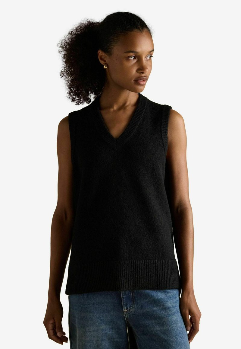 Czarny bezrękawnik z dzianiny z dekoltem w serek, z teksturą prążkowaną na dole. Połączony z jasnoniebieskimi dżinsami.